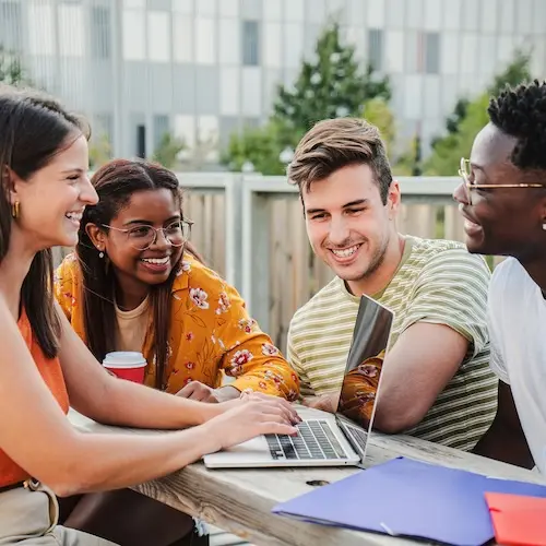 Four young adults are gathered around a laptop at an outdoor table, engaged in conversation and smiling. They appear to be collaborating or studying together, with a colorful folder and coffee cup present on the table. The setting suggests a casual, friendly atmosphere.