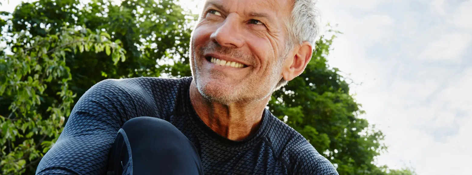 A smiling older man, dressed in a black athletic shirt, leans against his knee while enjoying a moment outdoors. The background features lush green foliage and a partly cloudy sky, suggesting a bright and cheerful atmosphere.