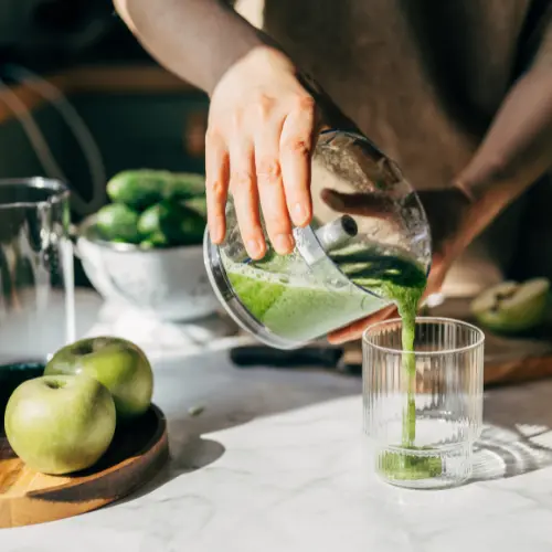 A person pours green smoothie into a clear glass from a blender. Fresh green apples and cucumbers are visible on a countertop. The scene suggests a healthy beverage preparation.