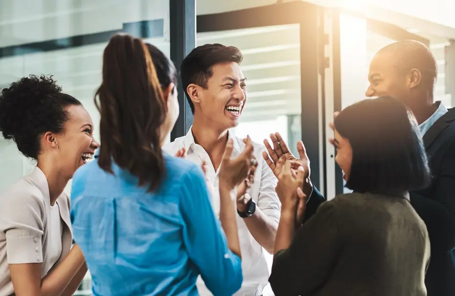 مجموعة من الأشخاص يتحدثون معًا في بيئة مهنية، يظهر عليهم السعادة والضحك. يتمتعون بلحظة من التفاعل الإيجابي، مع بعضهم البعض، بينما يجلسون في مكان ذو إضاءة جيدة.