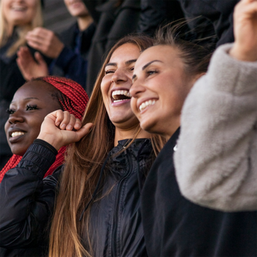 A group of three young women is smiling and cheering enthusiastically. They appear to be enjoying a lively event, with one woman raising her arm in excitement. The atmosphere conveys joy and celebration.