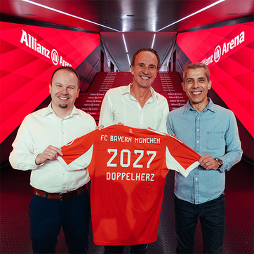 Three men stand together in a hallway at Allianz Arena, holding up a red FC Bayern München jersey featuring the year "2027" and "DOPPELHERZ." The background shows illuminated stairs leading to the arena, emphasizing the team's vibrant branding.