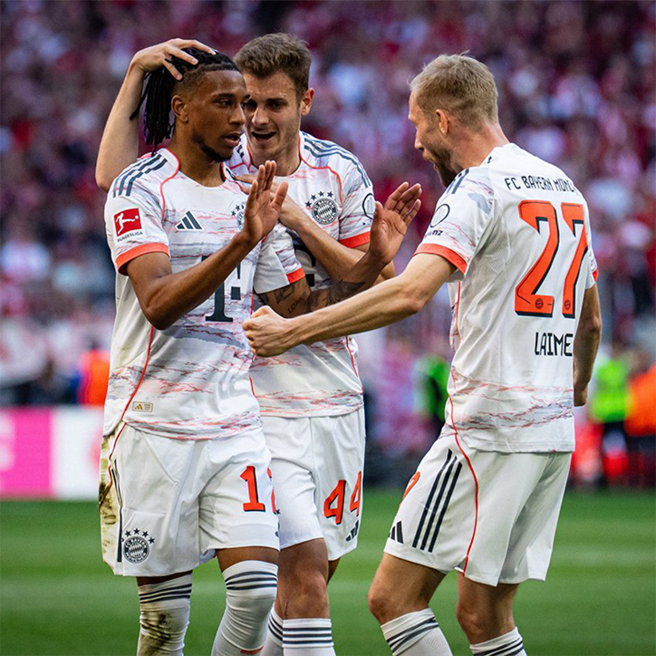 Three soccer players from Bayern Munich celebrate a goal. One player, wearing number 14, is in the center with teammates, smiling and raising hands in a gesture of joy. They wear white and red jerseys, with a blurred crowd in the background, indicating a lively match atmosphere.