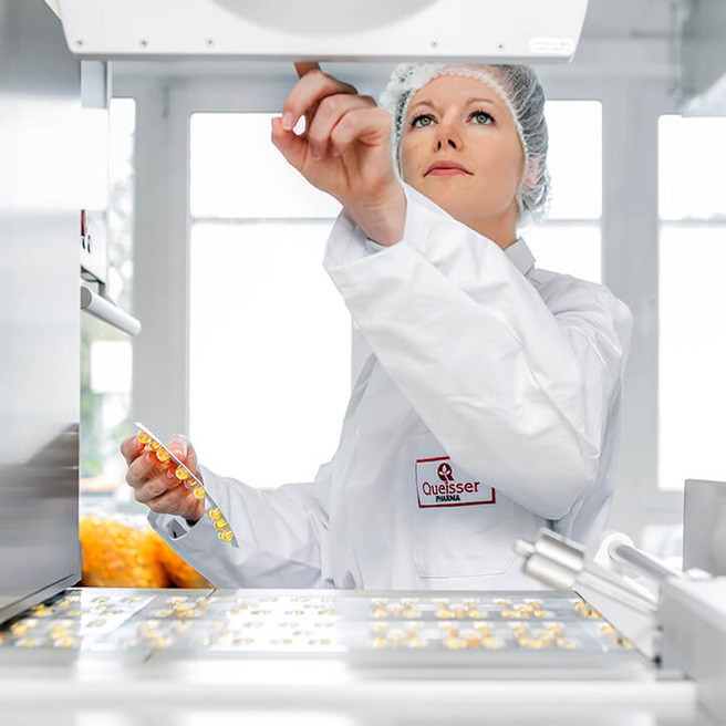 A woman in a lab coat and hairnet is inspecting small items on a conveyor. She holds a tray in her left hand while looking upward, focusing on a machine above. The setting appears to be a clean, professional food production facility.