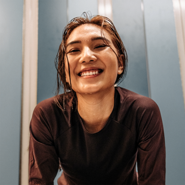A smiling young woman with wet hair leans forward, exuding joy and enthusiasm. She is wearing a dark athletic top, suggesting an active lifestyle. The background features light blue walls, creating a vibrant atmosphere. The image conveys a sense of happiness and accomplishment.