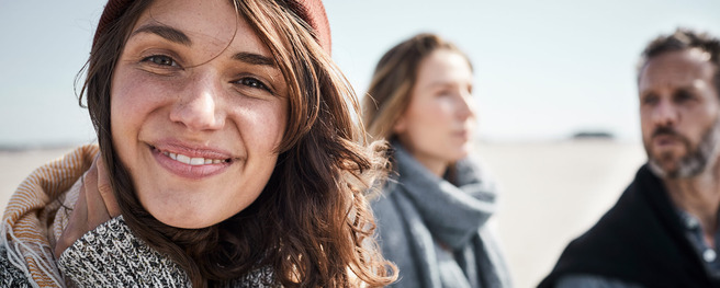 A smiling woman in a cozy sweater and beanie is in focus, with two blurred figures, one man and one woman, slightly behind her. The scene suggests a relaxed outdoor setting, possibly on a beach. The sunlight enhances the warmth of the moment.