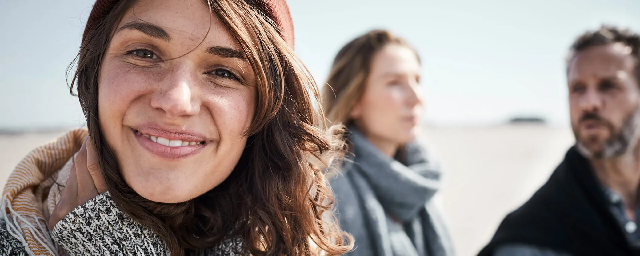 A smiling woman in a cozy sweater and beanie is in focus, with two blurred figures, one man and one woman, slightly behind her. The scene suggests a relaxed outdoor setting, possibly on a beach. The sunlight enhances the warmth of the moment.